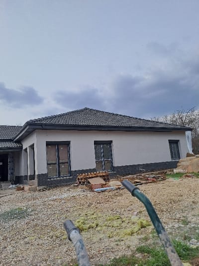 The image presents a construction site in the city of Kamenets, Bulgaria, under a predominantly overcast sky. A single-story residential building, appearing to be under active construction, is centrally positioned. The building's exterior features light grey stucco above a darker grey, stone-effect foundation finish. It is topped by a dark grey tiled, gabled roof with a dark rain gutter running along the eaves. The visible facade contains two large, dark-framed openings with interior protective coverings or film, likely intended for doors or large windows, and one smaller, similarly framed and covered window. An additional open entryway on the left side of the structure reveals interior construction elements. The ground in the foreground and midground consists primarily of gravel and exposed earth, with scattered patches of green vegetation and yellow-green fibrous insulation material. Construction debris, including stacked wooden pallets, planks, and loose red bricks, is concentrated near the building's base. In the immediate foreground, two dirt-smudged metal handles, consistent with those of a wheelbarrow, are partially visible and angled towards the viewer. Bare tree branches are discernible in the distant background on the right. No individuals are present in the scene.