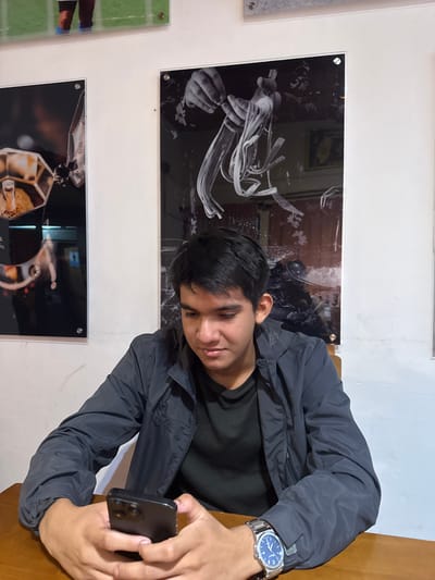 A person is holding a smartphone in front of a wall with photographs on display, likely in an indoor space in Chipilo de Francisco Javier Mina (Chipilo), Mexico. The individual is wearing a dark-colored jacket and a watch is visible on their wrist. There are at least three framed photographs on the wall. One appears to show a hand manipulating noodle-like objects. Another shows a complex, possibly machine-related, object, with the lowest photograph showing feet on a green background. The person is likely taking a photo of the images on the wall.