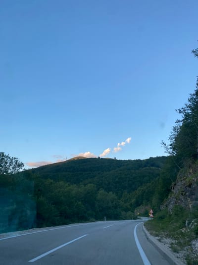 The image depicts a winding asphalt road passing through a lush, mountainous landscape in the vicinity of Praga, Montenegro. The road is marked with white lines and has a concrete barrier along the outer edge. Steep, tree-covered hills rise on both sides of the road. On the right side, a rocky cliff face is visible, with sparse vegetation growing on it. In the distance, a mountain summit is crowned with what appears to be a communication tower. The sky is a clear blue with a few scattered white clouds. Traffic signs, including one indicating a series of curves, are visible along the roadside. The overall scene suggests a rural, mountainous environment with a well-maintained road suitable for travel. There are no people or vehicles visible in the image.
