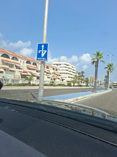 The image shows a street scene in La Manga Del Mar Menor, Spain. A blue and white sign on a metal pole indicates "HOTEL" with a directional arrow pointing straight ahead and a left turn. The buildings in the background appear to be apartments or hotels with white facades and orange tiled roofs. Palm trees line the street, and the sky is blue with scattered clouds. The perspective suggests the photo was taken from inside a vehicle, as the dashboard is visible in the foreground.