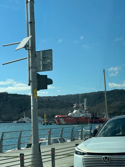 A daytime image, stated to be located in Sultangazi, Turkey, depicts a vibrant waterfront scene under a clear blue sky with sparse clouds. In the left foreground, a grey utility pole stands vertically, equipped with a square solar panel, two horizontal metal bars, and the rear of a rectangular street sign. A dark traffic signal is mounted on the pole, which also displays a yellow warning sticker reading "DANGER / TEHLİKE." A grey metal railing runs horizontally across the lower frame, separating the street from the water. A small white boat is partially visible docked behind this railing. On the right foreground, the front section of a white Honda HRV, including its grille and part of a headlight, is visible, with a faint reflection of a person in the windshield.

The midground features a body of water with visible surface ripples. A large, red and white vessel, identified as "Kıyı Emniyeti" (Coastal Safety) and "COASTAL SAFETY," is centrally positioned on the water. This vessel possesses a multi-level superstructure, a mast, and deck equipment. To its left, a yellow buoy or docking structure is present, alongside at least two smaller white vessels further back. A tall, light-colored mast of another sailboat is visible to the right of the red vessel.

The background comprises treed hills rising from the opposite shoreline. A small bird is observed in flight against the clear blue sky, above the sailboat mast. The scene is brightly illuminated by natural daylight.
