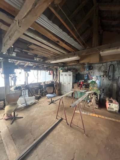The image depicts the interior of a functional, well-used workshop located in Podaiva, Bulgaria. No individuals are present.
The workshop features a dirt or concrete floor, uneven and marked with patches of dust and debris. The walls are constructed of rough stone or concrete blocks, painted a faded light gray, with visible staining and discoloration. The ceiling exposes wooden beams and rafters, with sections of corrugated metal sheeting and wooden planks forming the roof structure above. Several fluorescent light fixtures are mounted to the ceiling beams, providing artificial illumination in addition to natural light.
Along the far wall, large windows admit bright, diffused light, obscuring the exterior view. A heavy-duty drill press is positioned near the left window, on a metal workbench. To the right of this, a blue cushioned swivel chair is visible. Further right, another workbench holds various tools and equipment, including what appears to be an anvil and a green patterned cloth draped over a piece of machinery.
On the left side of the workshop, two gray, multi-compartment metal lockers are situated against the wall. Above these, shelves and hooks hold various items including small boxes and bags. The back wall, to the right of the windows, displays an array of hanging items including coiled electrical cables, tools, and plastic bags. A light-colored, possibly white, face mask or protective gear is also hanging.
In the mid-ground, two red-painted metal trestles (sawhorses) with metal crossbars are positioned. A long, dark metal bar or beam lies on the floor, extending from the bottom left corner towards the center. In the far right background, a red and white portable generator or welding machine is visible on the floor. A light brown upholstered chair, possibly a swivel office chair, is in the left foreground. The overall environment suggests active use for fabrication or repair work, characterized by a utilitarian layout and accumulation of tools and materials.
