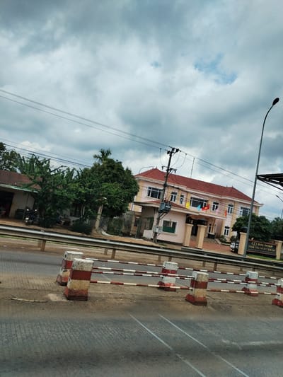 The image depicts a street scene in Ia Bang, Vietnam. A two-story building with a red tile roof and tan facade stands to the right, adorned with several flags. The building is identified by a sign as belonging to the DLGL Group, specifically the Gia Lai toll station. A row of red-and-white striped bollards and a metal guardrail separate the building from the road. Across the road, a modest building with a corrugated metal roof is visible, partially obscured by trees. Power lines stretch across the cloudy sky. A streetlight stands tall on the right side of the frame. The foreground shows the asphalt road surface.