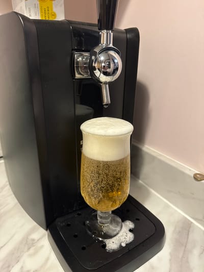 This scene is located in the city of Babergh, United Kingdom. The image displays a close-up of a black tabletop beer dispenser with a chrome tap mechanism. A stemmed clear glass, containing a golden, carbonated liquid and a significant white foam head, is positioned on the dispenser's black drip tray. Foamy white liquid has spilled from the glass onto the drip tray. The dispenser rests on a light-colored, possibly marble or laminate, countertop, against a plain, light pink wall. A partially visible white cardboard box with yellow text, indicating "ORIGINAL INDIAN TONIC WATER," is situated on top of the dispenser to the left. A small, circular, copper-colored object, consistent with a coin, is visible on the countertop to the right. No individuals are depicted.