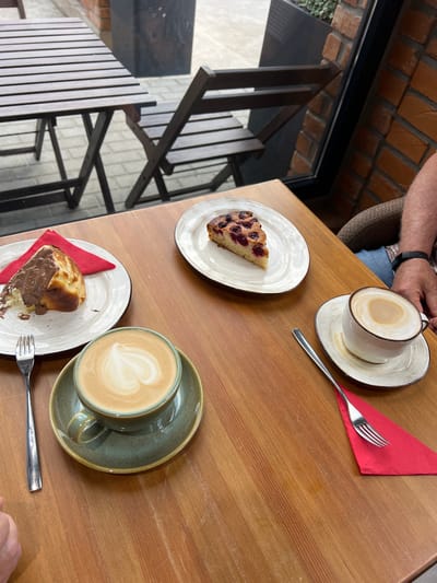 The image shows a top-down view of a wooden table at a cafe in Adzhiryuara, Abkhazia. Two plates with slices of cake and two cups of coffee are arranged on the table. One slice of cake appears to be a berry cake, while the other is covered in chocolate sauce. Both coffees are lattes with foam art. A red napkin and a fork are placed near one of the plates. The table is set against a backdrop of brick walls and dark wooden outdoor furniture, suggesting an outdoor seating area. The lighting suggests it is daytime. The scene depicts a casual moment of enjoying coffee and cake. There is no visible text in the image.