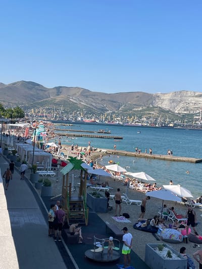 The image depicts a bustling beach scene under a clear blue sky, suggesting a warm, sunny day. A multitude of people are engaged in various beach activities. Some are sunbathing on lounge chairs, others are swimming or wading in the calm blue water, and many are walking along the pebble beach. Beach umbrellas provide shade for some of the sunbathers.
In the background, a large mountain range rises majestically, with a town or city built into its slopes, characterized by numerous buildings and industrial cranes, hinting at a port or industrial area. The sea is dotted with several small boats. The scene appears to be a popular tourist destination, given the density of people enjoying the recreational facilities. The overall atmosphere seems relaxed and cheerful, with people enjoying a day at the seaside. There is no visible text in the image. The rotation of the image suggests it was taken vertically.