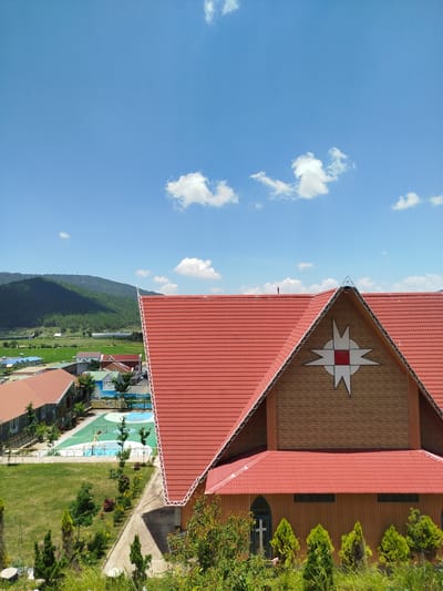 The image is a high-angle shot of a red-roofed building with a distinctive star emblem on its upper facade. The building is set against a backdrop of lush green hills and a bright blue sky dotted with white clouds. In the foreground, a vibrant green lawn is interspersed with trees and plants. To the left of the main building, there are other structures with red roofs, some appearing residential or part of a complex, and a visible basketball court with green and blue surfacing. The scene suggests a daytime view in a semi-urban or rural setting. The image is devoid of people.