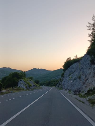 The image depicts a highway curving through a mountainous landscape near Vrela, Montenegro. The sky is a soft gradient of pale blue and orange, suggesting either dawn or dusk. On the right side of the road, a steep, rocky cliff face is covered in patches of green vegetation and trees. The road itself is asphalt, with clear white lane markings. A metal guardrail is visible on the left side of the road. In the distance, rolling green hills are silhouetted against the sky. There are no people or vehicles visible in the image. The scene is tranquil and natural, emphasizing the rugged beauty of the Montenegrin countryside.
