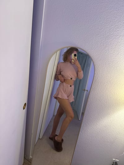 A young woman with blonde hair is reflected in a full-length mirror, wearing a pink ribbed long-sleeved crop top and matching ruffled shorts. She is holding a smartphone in her right hand to take the selfie. She is wearing brown fuzzy slippers. The mirror is positioned against a textured purple wall. To the left of the mirror is a white door with a gold handle. The floor is carpeted in a light beige color. The image appears to be taken indoors, in what could be a bedroom or dressing area. The context of Tustin, California, USA is not discernible from the visual information provided in the image.