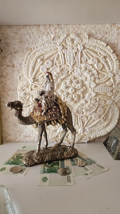 The image displays a decorative figurine of a man riding a camel, positioned on a surface covered with Russian banknotes and coins. The figurine is detailed, with the man wearing traditional Middle Eastern attire and the camel adorned with an ornate saddle blanket. Behind the figurine, a textured, circular decorative panel with intricate patterns and small pearl-like embellishments is visible, mounted on a textured wall. To the right of the central display, a small, metallic pyramid-shaped object is partially visible. The banknotes appear to be of low denomination, and the coins are silver-colored. The overall scene is a still-life arrangement, likely indoors, with a focus on decorative items and currency. There is no identifiable location in Lubnya, Russia, depicted or suggested by the elements within the image.