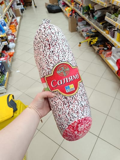 The image shows a person holding a large, red and white cylindrical pillow with a label that reads "Salami." The label is in Cyrillic, and the image shows the blue and yellow colors of the Ukrainian flag. The person's arm and wrist are visible, and they are wearing a silver bracelet. The background shows a supermarket or retail store. The flooring is white tiled, and the shelves are stocked with various products. This scene is in a store located in Berehove, Ukraine.