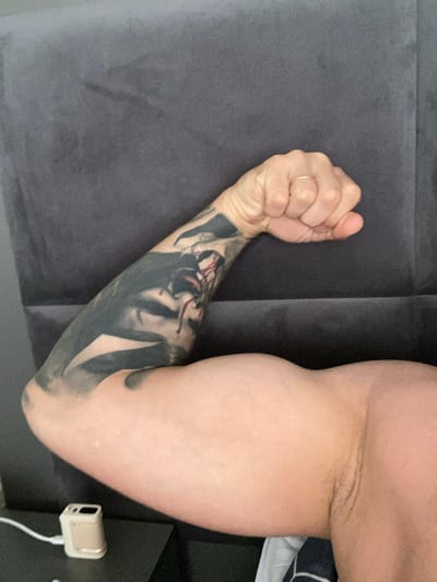 The image shows a close-up of a person's flexed bicep and forearm, which has a large, dark tattoo extending from the wrist up the arm. The bicep is well-defined. The person is wearing a gold ring on their right hand, which is clenched into a fist. The background is a dark grey upholstered surface, possibly a headboard or sofa. A small, light-colored electronic device with a white cord is visible on a dark surface in the lower left corner. There is no information in the image to suggest it is located in Chalda, Russia.
