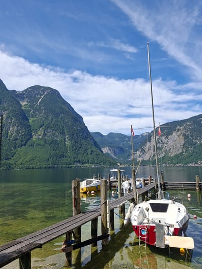 This is a medium-shot, eye-level, full-color photograph taken outdoors on a bright, sunny day in Obertraun, Austria, likely on Lake Hallstatt. The foreground is dominated by a wooden pier, with two boats moored to it. The closest boat is a white and red sailboat, tilted on its side, with its stern facing the viewer. It has a red hull and white superstructure, with fenders hanging over its sides. A small ladder is attached to its stern. To the left of this boat, a yellow boat is visible, also moored to the pier. Further in the background, several other boats are docked. The water is clear, revealing the rocky lakebed. Behind the lake and pier, a densely forested, steep mountain range rises majestically. The sky above is a vibrant blue with scattered white, wispy clouds. The scene depicts a tranquil lakeside setting with moored sailboats and a dramatic mountain backdrop.