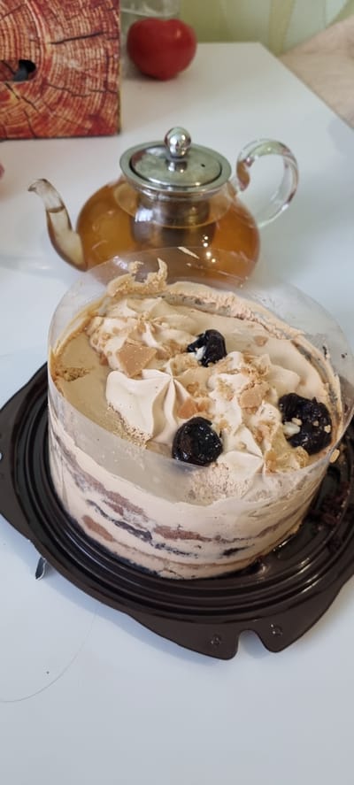 The image displays a close-up, top-down view of a layered cake and a glass teapot on a white surface. The cake is adorned with whipped cream, dark cherries, and crumbled biscuit pieces. It sits within a dark brown plastic container. To the left of the cake, a clear glass teapot filled with amber liquid is visible. A red, round object, possibly a tomato or a decorative item, is also present to the left of the teapot, near a wooden object with a prominent wood grain pattern. The lighting suggests it is daytime, and the overall scene appears to be indoors, possibly in a home or cafe setting in Romashkovo, Russia. There are no people visible, and no text is present in the image. The focus is on the food items and their presentation.
