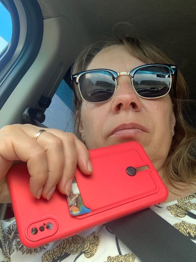 A woman in sunglasses is taking a selfie in a car. She is holding a red phone case with a card slot. The reflection in her sunglasses shows the road and trees. The car's interior is visible, including the seatbelt. The background is out of focus. The image appears to be taken during the daytime. There is no discernible information in the image to determine the specific location within Грозаўскі сельскі Савет, Belarus.