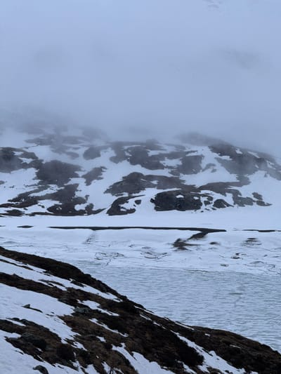 The image presents a serene, snow-covered alpine landscape, consistent with the specified location of Poschiavo, Switzerland. The scene is dominated by rugged mountains, a body of water, and pervasive fog.

In the foreground and extending into the midground on the right, a steep mountain slope descends, characterized by dark, exposed rock formations interspersed with significant patches of white snow and some sparse, brown, wintry vegetation. This creates a textured pattern of light and shadow. Adjacent to this slope, a body of water, likely a lake, stretches across the midground. Its surface appears partially frozen or covered in fragmented ice, showing intricate patterns, reflecting the cold conditions.

Further into the background and to the left, another large mountain face rises. This peak is heavily covered in snow, with darker rocky sections peeking through. A substantial portion of this mountain, particularly its upper reaches, is enveloped in thick, greyish-white fog or low-hanging clouds, which softly obscure the details and create a sense of vastness and mystery. The fog diffuses the light, contributing to the muted, monochromatic palette of the scene.

The overall lighting suggests an overcast daytime, providing soft, even illumination without harsh shadows. The weather is clearly cold, characterized by snow and fog, indicating either a deep winter or early spring season. There are no people, animals, or man-made structures visible, emphasizing the untouched natural environment. No text is present in the image. The scene evokes feelings of tranquility, cold, and the stark beauty of a mountainous wilderness.
