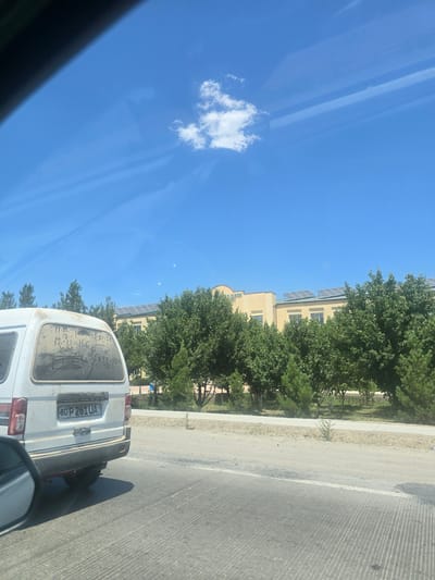The image captures a view from inside a vehicle looking out at a street scene in the city of Yukari-Gazbiran, Uzbekistan. In the foreground, the rear of a white, somewhat dirty van is visible, with a license plate reading "80 P 261 UA". Writing, possibly mathematical equations, is scrawled on the van's rear window. Beyond the van, a row of green trees lines the street. Further in the background, a light-colored building with solar panels on its roof is situated amongst the trees. The sky is clear and blue with a few scattered clouds, and the sun appears to be shining. The road surface in the foreground shows parallel lines of shadow.