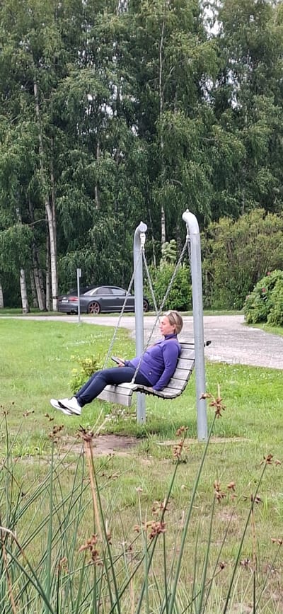 In Kohatu, Estonia, a woman is sitting on a wooden swing, holding a mobile phone. She is wearing a purple jacket and dark pants with white sneakers. The swing is suspended by metal chains attached to two grey metal poles. The background features a line of trees with green foliage and a grey car parked behind them. The ground is covered in green grass and tall, thin reeds in the foreground.