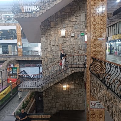 An indoor shopping mall features a multi-level design with stone walls and ornate metal railings. A woman stands on a staircase, with a red bag, between levels. Signage for stores is visible, including "Modelos," "PARA EL HOGAR" and "LORD PIE". A "No Smoking" sign is mounted to a tile-covered pillar. A man is sitting on a bench near the bottom of the image. Balconies and walkways crisscross the space, creating a layered perspective.