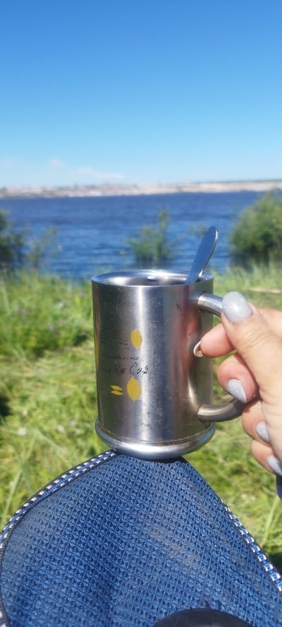A first-person perspective shot shows a hand holding a metal mug with a spoon inside. The mug has two yellow lemon-shaped designs on it. The background is a body of water with a distant shoreline under a clear blue sky. The foreground shows a blue textured fabric, possibly clothing. The scene is outdoors, with green grass and some plants visible. The location is identified as Mokhsogollokh, Russia, suggesting a natural or recreational setting.