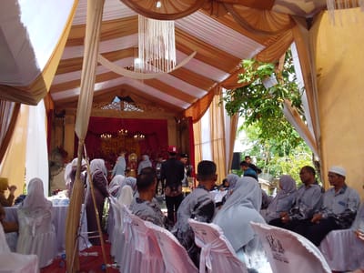 This media file depicts an outdoor or semi-outdoor event taking place under a large, decorated tent in Gampongjawa, Indonesia, during daytime. The tent's ceiling is draped with alternating stripes of white and golden-brown fabric, and a decorative white tassel chandelier hangs centrally.
Numerous individuals are present, predominantly seated on white-covered chairs arranged in rows. Many women wear hijabs in various colors like light grey, white, and brown. Several men are dressed in patterned shirts, possibly batik, and one man on the right wears a white peci (skullcap). A man in a red cap is seen standing near the front of the tent.
At the far end, a decorated stage area is visible with a rich red velvet backdrop, adorned with ornate gold-colored carvings and chandeliers. Other objects include tables with white tablecloths, a standing fan, and sound system speakers. Lush green foliage and trees are visible outside the tent, indicating the event's outdoor setting and bright natural light.
The text "EVA DECORATION 0852 3157 3686" is visible on the white covers of several chairs. Another chair cover has the numbers "88 2 855" faintly printed. The scene suggests a formal gathering or celebration, such as a wedding or reception, given the elaborate decorations and attire of the attendees.