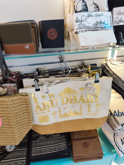 The image shows a retail display of souvenirs and merchandise in Al Matar, United Arab Emirates. The merchandise includes several wallets, tote bags, and folded fabrics, all featuring designs and text related to Abu Dhabi. Some of the items are displayed on shelves, while others are hung on hangers. The color palette is dominated by neutral tones like brown, black, and white, with accents of gold on the tote bags. There are no people visible in the image. The environment appears to be an indoor retail space, possibly a gift shop or souvenir store.