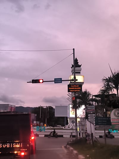 The image depicts a street scene at an intersection in Lamprehlam Teungoh, Indonesia, likely during dusk or dawn, given the cloudy, purplish-pink sky.
In the foreground, the rear of a black truck is visible, with the brand "SUBISHI" and model "COLT" clearly legible, and its tail lights illuminated. To the left, a red traffic light is active, signifying a stop. Attached to the traffic light pole is a blue sign in Indonesian stating "DILARANG MENEROBOS" (Forbidden to run a red light).
To the right, a multi-tiered, pagoda-like tower structure stands, decorated with various illuminated signs. A prominent orange LED sign on the tower displays the message "TAATILAH RAMBU-RAMBU LALU LINTAS" (Obey traffic signs). Surrounding the tower and along the roadside are numerous other signs. These include advertisements for "GRAHA MECCA Percetakan" (Printing), "SPANDUK" (Banners), "X BENNER" (X-Banners), and "CETAK BUKU" (Book Printing). Other signs mention "CPNS," various educational institutions like "DAYAH AWJA AL UL INGIN JRY QURBAN," "SMA IT AWJA," and "SMP IT AWJA," as well as a location "DESA KAYEE KUNYET KEC. BLANG BINTANG KAB, ACEH B." A sign with "HARGA MULA" and a phone number "05214" is also visible.
The road is paved, with motorbikes partially visible, indicating active traffic. Palm trees and other vegetation are present on the right side of the road. In the background, buildings and distant mountains are discernible. A large blue "BSI" sign is partly visible. Power lines crisscross the upper portion of the frame against the cloudy sky.
