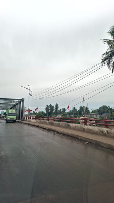 The image captures a view from a vehicle traveling on a wet road towards a bridge. On the left, a green truck is visible on the bridge. The bridge itself has a metal truss structure and is lined with red railings. Several Indonesian flags are seen on the bridge. The background features lush greenery with palm trees and some buildings. Utility poles and overhead power lines are also present. The sky is overcast, suggesting a cloudy day. The scene appears to be in a tropical or subtropical environment. The presence of the Indonesian flags suggests a location within Indonesia.