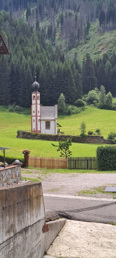 The image shows the iconic St. Magdalena Church in Santa Maddalena, South Tyrol, Italy. The small, white church with red trim and a dark onion dome is situated on a vibrant green hillside, bordered by a low stone wall. Behind the church, a dense, dark green coniferous forest blankets a steep mountain slope, with patches of lighter green grass and trees interspersed on the terrain. The sky is overcast, casting a soft light on the scene. In the foreground, a light-colored gravel driveway meets a darker paved area. To the left, a concrete structure is visible, along with a stone wall adorned with a flower pot and a wooden sign. A rustic wooden fence runs across the mid-ground, separating the driveway area from the church's property. Two figures are standing near the church. The overall atmosphere is serene and picturesque, characteristic of the Italian Alps.