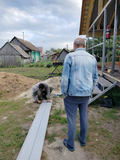 In Вазерки, Russia, two men are working outdoors near a partially constructed building. One man, wearing a blue denim jacket and grey sweatpants, stands facing away from the camera, observing the work. The other man, dressed in a grey sweatshirt and dark pants, kneels on the ground, measuring and marking a light grey siding panel with a ruler and what appears to be a utility knife. The siding is laid out on the ground. In the background, several weathered wooden structures, a pile of sand, and overgrown grass are visible. A metal frame for a building extension is also present, with a red fire extinguisher attached. The sky is overcast.