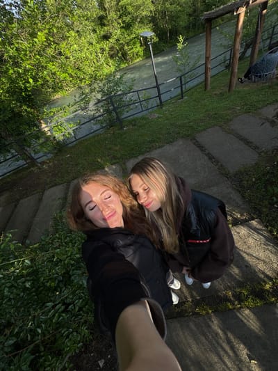 Two young women are posing closely together for a selfie in an outdoor setting. The woman taking the photo has red hair and is wearing a black jacket. The other woman has blonde hair and is wearing a brown hoodie and a black puffer jacket, which has a red label on it. They are standing on a path made of concrete blocks, with grass and greenery visible on either side. A fenced waterway and additional trees are also visible in the background. The location appears to be outdoors, possibly a park or garden. The city and specific location cannot be determined from the visual information provided.