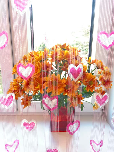 A vase of orange chrysanthemums sits on a windowsill, framed by a beaded curtain with pink and white hearts. The scene is indoors, near a window that looks out onto trees. The flowers are vibrant, with many petals. The beaded curtain is decorative, with a repeating pattern of hearts.