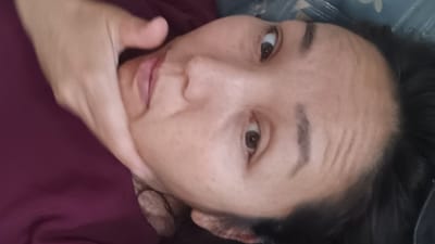 The image is a close-up, sideways, and slightly blurry portrait of a person's face. The person's left hand is visible, touching their chin. Their eyes are open and looking upwards, and their lips are slightly parted. The person is wearing a dark maroon shirt. The background appears to be a textured surface, possibly a bed or a mattress, with some light-colored patterns. The lighting is moderate, with shadows emphasizing the contours of the face. No specific location in Құрманғазы ауылдық округі, Kazakhstan, can be identified from the image.