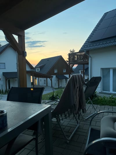 The image depicts an outdoor seating area at dusk, likely in Milná, Czechia. A dark-colored table with an ashtray is in the foreground, with two chairs partially visible. One chair is draped with a light brown blanket. In the background, several modern houses with dark roofs and white walls are visible. Some houses feature solar panels. The sky shows a gradient of blues and oranges, indicating sunset. The overall setting suggests a residential area with a tranquil, early evening atmosphere.