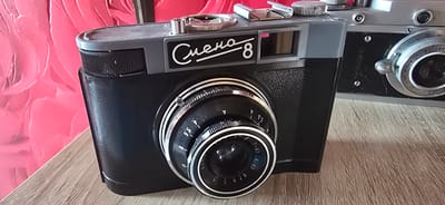 This media file features two vintage film cameras, likely in a domestic setting in Gostagayevskaya, Russia. The primary subject is a black and gray "Smena 8" camera, positioned in the foreground. It has a distinctive silver lens assembly with aperture markings visible, including "5.6" and "8". The camera also has a silver winding knob and a chrome viewfinder. The "Smena 8" text is prominently displayed in white cursive script on the camera's body.
To the right and slightly behind the Smena 8 is another vintage camera, appearing to be silver and black. Some text is visible on its lens assembly, including "N80384" and "F=5см".
The cameras are placed on a wooden surface, possibly a table, which has a light brown, grainy texture. The background features a vibrant, textured pink surface with a floral or petal-like pattern. The lighting suggests an indoor setting, possibly during the daytime, with no specific weather cues. The overall impression is one of a still-life composition focusing on retro photography equipment.