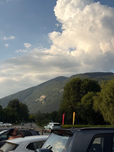 This image depicts a scenic mountain landscape with a parking lot filled with cars. The sky is partly cloudy, with large, white cumulus clouds dominating the upper left portion of the frame, set against a clear blue sky. A lush green mountainside covered in dense trees stretches across the right side of the image, with some lighter rocky patches visible on its slopes. A road winds its way up the mountain in the middle ground, with several cars parked along its edge. In the foreground, a white car is parked, and a young woman with dark hair is leaning out of the window, touching her hair. She is wearing a white top. Her expression is relaxed. Another car, a dark SUV, is parked behind her. Two colorful flags, one yellow and one red, are planted in the grass near the parked cars. The yellow flag has text that appears to be "DISC GOLF" and the red flag has text that appears to be "DISC". The image appears to have been taken during the daytime, possibly late afternoon, given the soft light illuminating the clouds and the shadows cast by the trees. The overall atmosphere is peaceful and suggests a recreational outing in a natural setting. The location context provided is Novalaise, France.