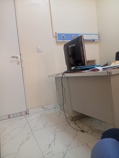 The scene depicts an interior room in Okengwe - Obehira, Nigeria, featuring light-colored walls and a white tiled floor with grey marble-like veining. A white door with a silver handle and round lock is partially visible on the left, showing a red and white 'SST ALUMINIUM PRODUCT' label at its lower edge. Mounted on the wall is a blue and white medical service panel, equipped with two circular ports (likely for medical gases) and an electrical outlet. Below a visible electrical conduit on the wall, a light switch or outlet is present. A light-colored desk occupies the right side of the frame, upon which a black HP computer monitor is positioned, facing away from the viewer. To its right, a stack of folders and papers is visible. Black electrical cables from the monitor descend towards a white power strip located on the floor under the desk. A dark, possibly upholstered, object is partially visible at the extreme right edge of the frame. No individuals are present.