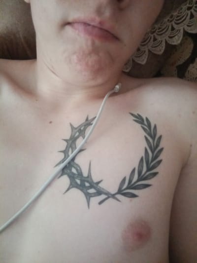 The image shows a close-up of a person's chest with a tattoo of a laurel wreath and thorns. A white cable, possibly from headphones, is visible across the chest. The person's neck and chin are also in view, showing some skin imperfections. The background is blurred and appears to be fabric, possibly a bedsheet or blanket, with a lacy pattern. The scene is not specific to Borodino, Russia, or any other particular city.