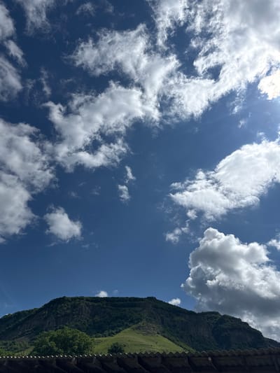 The image captures a scenic view of a lush green mountain under a bright blue sky dotted with fluffy white clouds. In the foreground, a portion of a rustic roof made of corrugated material is visible, suggesting a rural or semi-rural setting. The mountain itself is densely covered in trees, with some rocky outcrops visible on its steep slopes. The overall impression is one of natural beauty and tranquility. It is not possible to definitively identify the specific location within the city of Каменномостский, Russia based solely on this image.