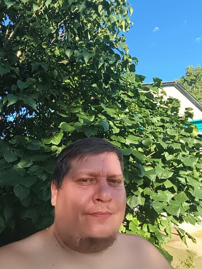 The image is a selfie taken by a man in front of a large, leafy green tree. The man is facing the camera and has dark hair, brown eyes, and a light complexion. He is shirtless and has a slight smile on his face. Behind him, the tree's foliage fills most of the frame, with some sky visible through the leaves. A portion of a white building with a dark roof is visible in the upper right corner of the image, along with a strip of turquoise siding. The overall scene suggests an outdoor setting on a sunny day. The image does not contain any specific landmarks or indicators that would allow for precise location identification within the city of Konstantinivka, Russia.
