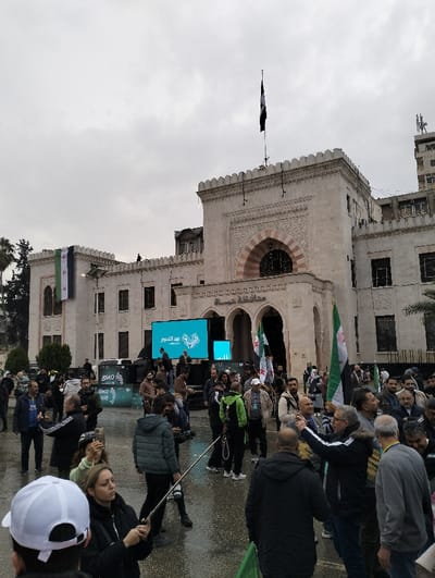A crowd gathers in front of a large, ornate building beneath a cloudy sky. Several people are holding Syrian flags. A tall screen displaying text in Arabic stands between the crowd and the building's entrance. The architecture of the building includes arches and decorative stonework, topped with a flagpole flying a black and white flag. The ground appears wet, suggesting recent rain.