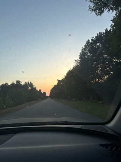 The image shows a view from inside a vehicle looking out through the windshield. The perspective is slightly angled, with the sky taking up the left portion of the frame and a road leading into the distance on the right. Dense trees line both sides of the road. The sky transitions from a clear, pale blue at the top to a soft orange and yellow hue near the horizon, indicating it is likely sunset. There are some small specks visible on the windshield. The interior of the car, including parts of the dashboard and A-pillar, is visible on the right side of the image, providing a dark, contrasting frame to the outdoor scene. The overall mood is serene, characteristic of a scenic drive at dusk. The crops suggest details of foliage and the car's interior structure. No people are visible. No text is present. The location context is Dovhe, Ukraine.