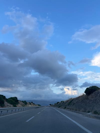A POV shot from a vehicle traveling on a paved road through a rural, hilly landscape under a cloudy sky. The road has white lane markings and a metal guardrail on the left. Vegetation, consisting of dry grass and shrubs, lines the sides of the road. In the distance, a white car is visible on the road. The sky is a mix of blue and grey clouds, with some areas of bright light suggesting sunlight breaking through. The scene appears to be located in a suburban or rural area, possibly on the outskirts of Manfredonia, Italy, given the Mediterranean vegetation and terrain. There are no people visible.