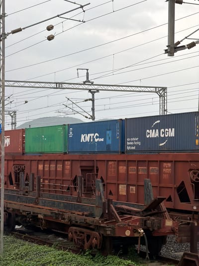 The image depicts a freight train carrying containers, parked on railway tracks in Saphale, India. The train consists of several open-top railcars loaded with shipping containers. In the foreground, a rusty, multi-level railcar is visible, with various metal components and what appears to be a coupling system. Behind this, three stacked shipping containers are prominently featured: a red container on the left, a green container in the middle, and a blue container on the right. The blue container bears the white logo and text "CMA CGM". Above the train, overhead power lines and supporting infrastructure for an electrified railway system are clearly visible against an overcast sky. The ground beneath the tracks is covered with lush green vegetation. No people are present in the frame. The scene appears to be an outdoor industrial or transportation hub, likely a railway yard or loading facility within Saphale.
