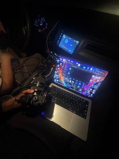 The image depicts a close-up, high-angle view of a person's hands operating a laptop and camera inside a vehicle at night. The laptop, a silver MacBook Pro, is open and displays a file explorer window filled with numerous colorful image thumbnails. Cables are connected to the laptop and a camera, which is being held by a person's left hand. The camera, a mirrorless model with a Tamron lens attached, has its lens pointed away from the viewer. To the left, another hand holds a smartphone displaying a time of 9:25. The vehicle's dashboard is visible in the background, with illuminated gauges and a car stereo displaying song information. The overall scene suggests a mobile workspace for content creation, likely photography or videography, within the city of El Paso, United States.
