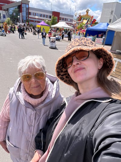 The image depicts two individuals, an older woman and a younger woman, in what appears to be an outdoor public area. The older woman has white hair and is wearing a light purple puffy jacket and glasses with round, gold-tinted lenses. The younger woman is wearing a leopard print bucket hat and round, brown-tinted glasses. The older woman is positioned in the lower part of the frame, while the younger woman is in the upper portion.

The background shows a cityscape with several tents and people. There are buildings with red and white facades, and blue and white tents are also seen in the distance. A large, colorful sculpture of a dragon-like figure is visible behind the tents. A crowd of people is milling around in the central area.

The image was taken in Valozhyn, Belarus.