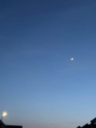 The image displays an outdoor scene in Easingwold, United Kingdom, captured during twilight hours. The majority of the frame is occupied by a clear sky, transitioning from a deep blue at the top to a lighter, pale blue-pink hue near the horizon. A distinct, pale crescent moon is visible in the upper right quadrant of the sky. In the lower left, a streetlamp with a bright, warm yellow illumination is present, its fixture and upper pole visible. Along the bottom edge of the frame, the dark, silhouetted rooftops of multiple buildings, consistent with residential or urban structures, are partially visible. No individuals, explicit actions, or interactions are discernible within the frame.