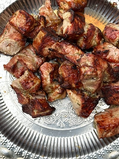 A close-up, overhead shot shows a metal platter filled with grilled meat. The meat pieces are irregular in shape and size, with a browned, slightly charred exterior and a pinkish-white interior. They are seasoned with herbs, visible as small green flecks. The platter itself is silver with a decorative, fluted rim and an embossed floral pattern. The meat appears to be arranged in a pile, some pieces overlapping others. The lighting suggests an outdoor setting, possibly a picnic or barbecue. The scene is devoid of people and activity, focusing solely on the presentation of the food. The location is identified as Podmoy, Russia, though no specific urban landmarks are visible to confirm this.