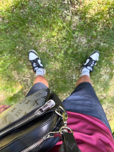 A photograph taken from a high-angle perspective shows a person standing on grass in an outdoor setting, inferred to be in Kanchevo, Bulgaria. The person is wearing dark grey shorts, black and white sneakers, and white socks. Part of a maroon garment is visible near the waist. A black Louis Vuitton bag is positioned to the right side of the frame, partially obscuring the person's body. The grass appears to be green and interspersed with small white flowers.