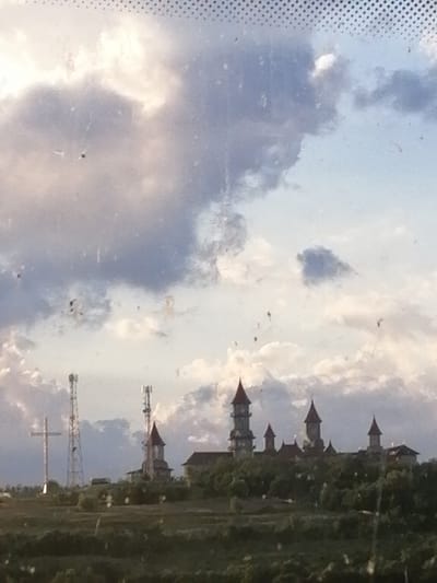 The image depicts a landscape with a sky full of clouds and a cluster of buildings in the distance. On the left side of the image, there are two tall communication towers and a large cross. In the center and to the right, a series of buildings with pointed roofs, reminiscent of a castle or a church complex, are visible. These structures are situated on a slightly elevated terrain covered with vegetation, including trees and bushes. The overall lighting suggests it is either sunrise or sunset. The scene appears to be an open area, possibly on the outskirts of a town or city. The location is identified as Roșiori, Romania.